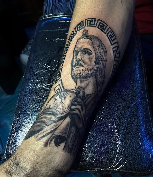 San Judas Tadeo en negro y gris sobre el antebrazo, con un marco de greca alrededor de la cabeza